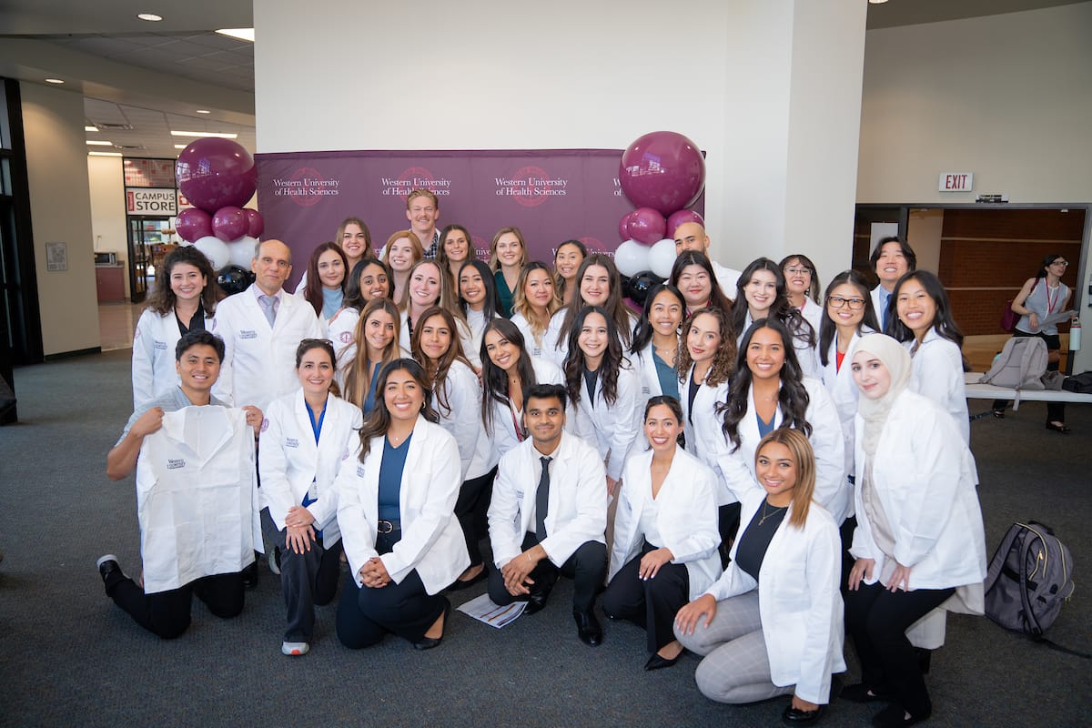 A group of students in white coats pose together and smile for a photo indoors, with balloons and a banner in the background.
