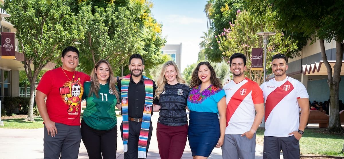 A group of seven people standing outdoors on a campus pathway, smiling at the camera. They are casually dressed and appear diverse in background. Trees and campus buildings are visible in the background.
