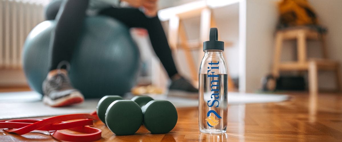 Close-up of a SamFit water bottle on a wooden floor next to two green dumbbells and a red resistance band. In the background, a person is sitting on an exercise ball.