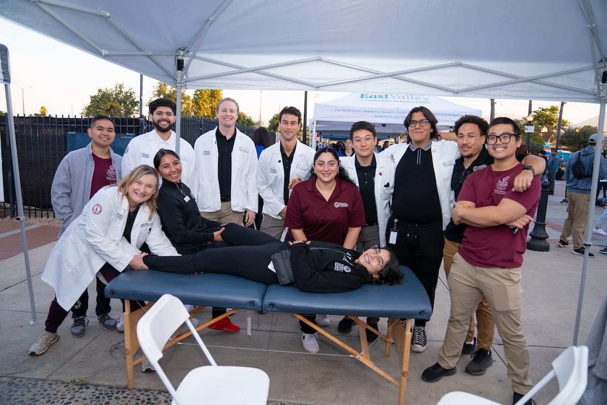 A group of people, some in lab coats and others in maroon shirts, stand and smile around a person lying on a massage table at an outdoor event.