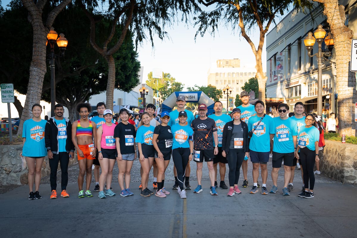 A group of runners gathers at a street race event, standing side by side in athletic wear and race bibs, with trees and a finish line banner in the background.