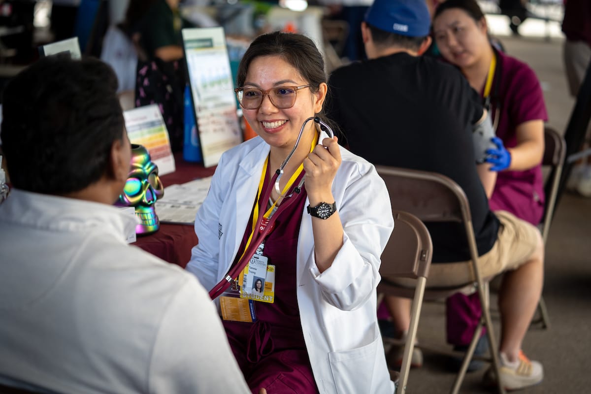 A female healthcare worker in a white coat holds a stethoscope and smiles while talking to a seated person at a community health event.