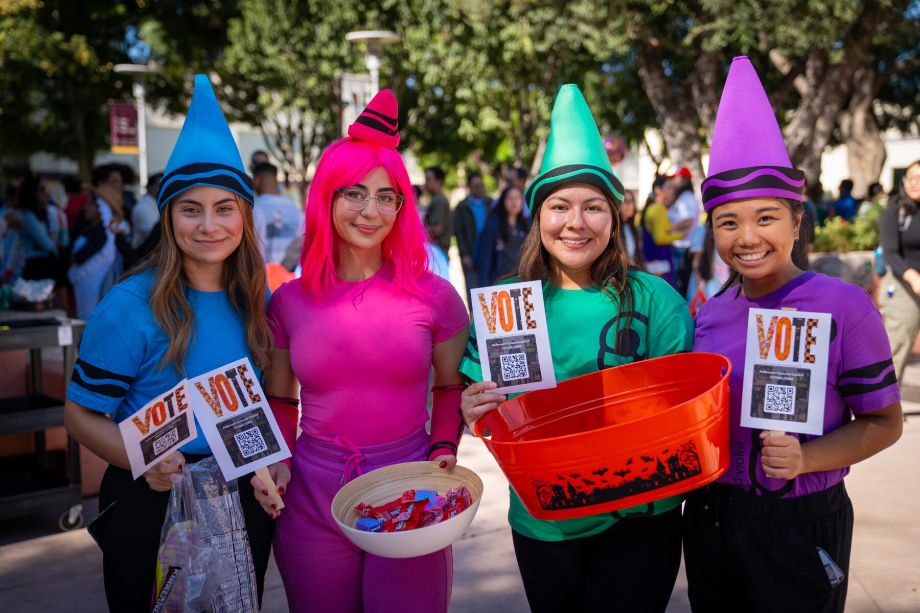 Four people in colorful costumes hold "VOTE" flyers with QR codes and baskets of candy at an outdoor event, with trees and other people in the background.