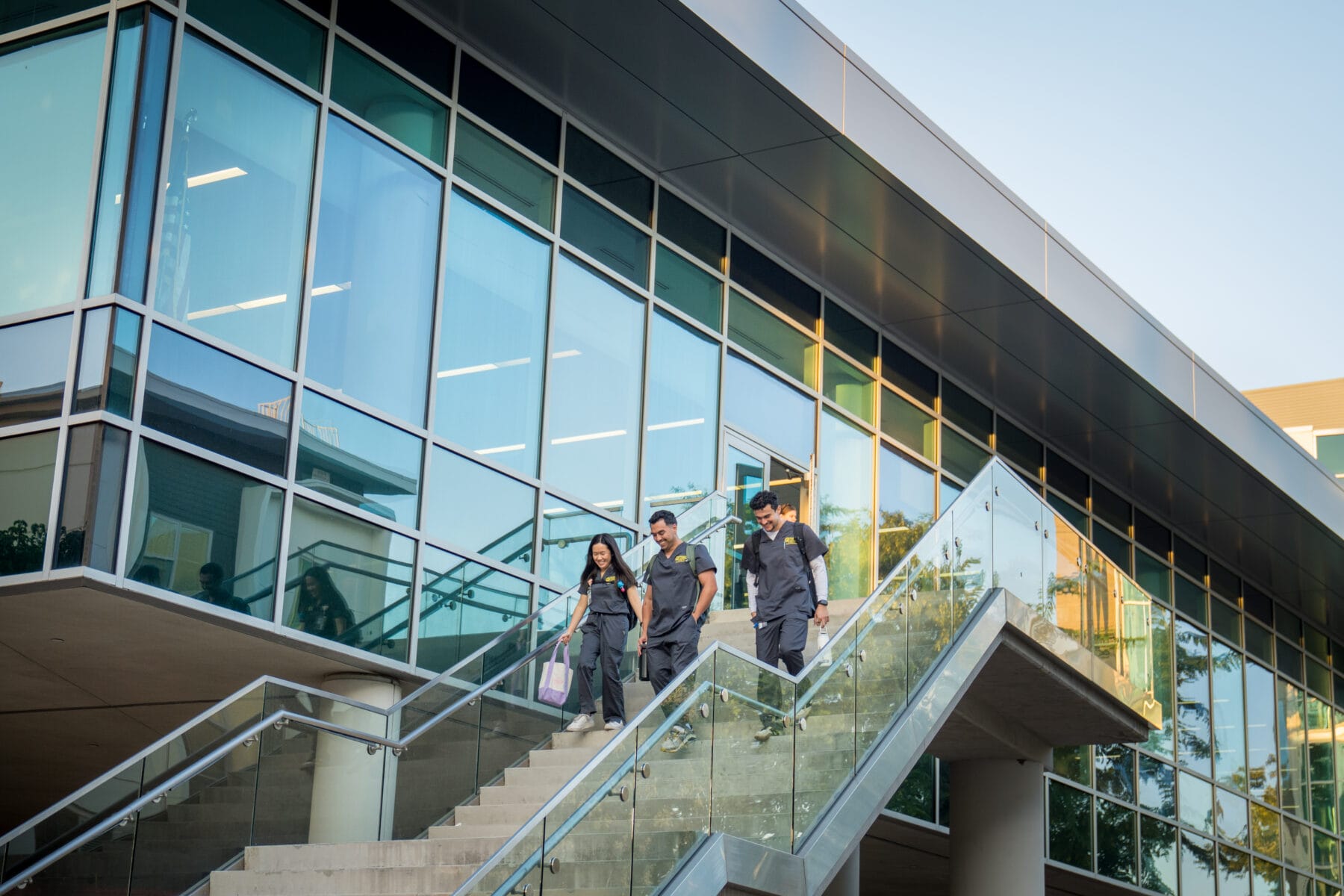Four people walk down exterior glass stairs beside a modern building with large windows on a clear day.