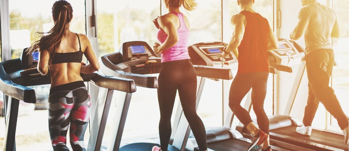 Four people running on treadmills in a gym, wearing athletic clothing. Sunlight streams through large windows in the background.