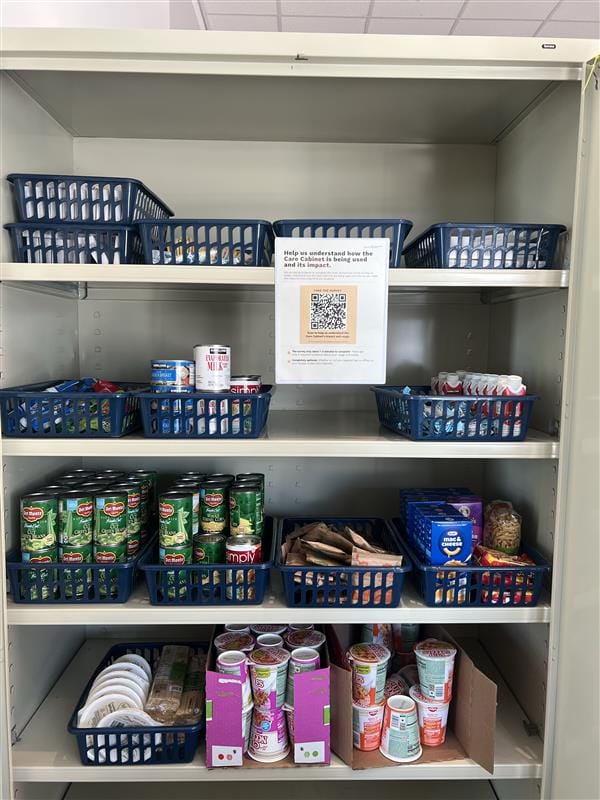 A metal shelving unit holds blue baskets filled with canned goods, packaged food, instant noodles, and small food items. A printed sign with a QR code is posted on the upper shelf.