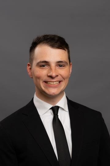 A person in a suit and tie smiles against a gray background.