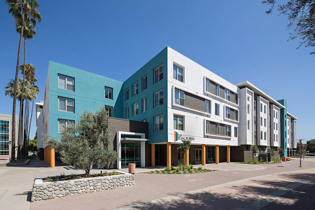 Modern four-story apartment building with teal and white exterior, large windows, and palm trees along a paved walkway under a clear blue sky.