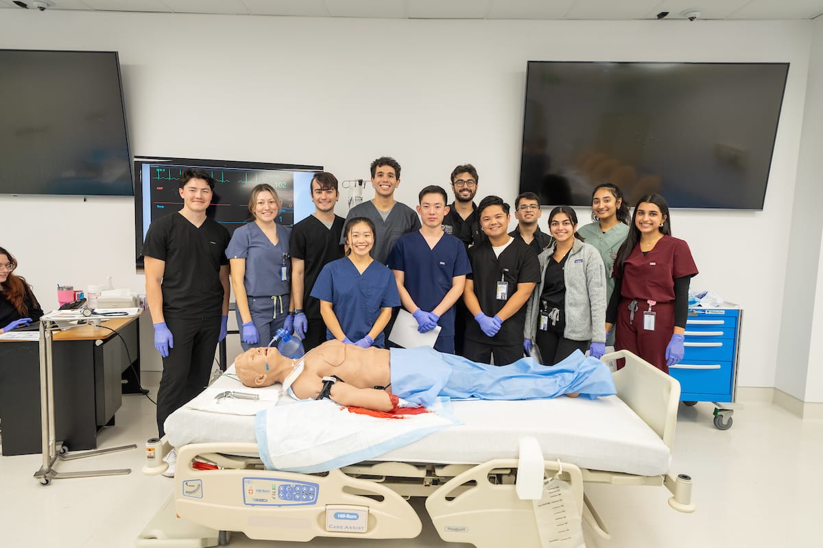 A group of healthcare professionals in scrubs poses around a medical bed with a lifelike human mannequin. The room has medical equipment and large monitors on the walls.