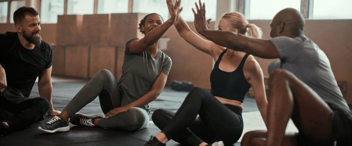 A group of four people, sitting on the floor in an exercise studio, are smiling and giving each other high-fives. Two men and two women are wearing workout clothes.