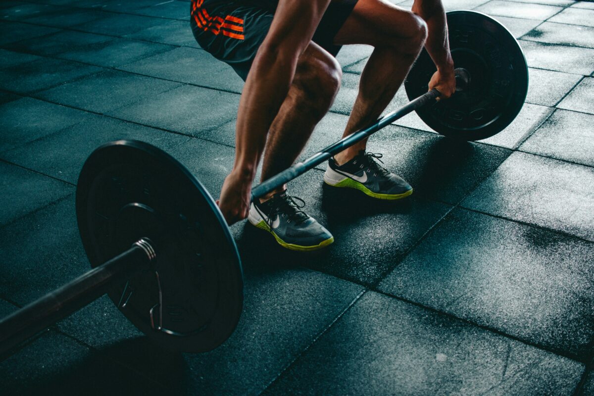 Man in athletic shorts and sneakers preparing to lift a barbell in a gym with a tiled floor.