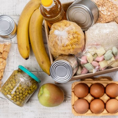 A variety of groceries including canned goods, bananas, pasta, oil, eggs, an apple, peas, crackers, and colorful marshmallows arranged on a white surface.