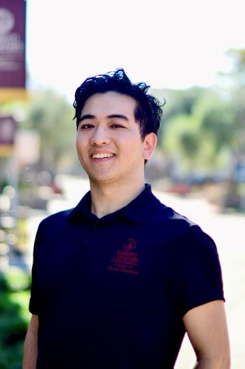 Smiling person in a dark polo shirt stands outdoors with blurred trees in the background.