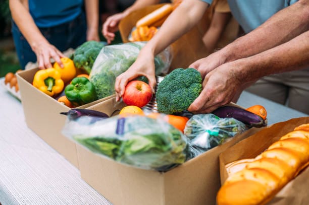 Several people pack cardboard boxes with fresh produce, including apples, broccoli, peppers, and bread, on a table.