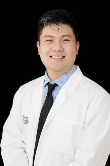 Person wearing a white lab coat over a striped shirt and tie, standing against a black background, smiling at the camera.