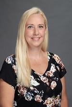 A woman with long blonde hair, wearing a floral top, smiles gently against a gray background.
