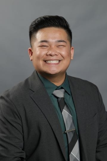 Person smiling broadly, wearing a dark suit, green shirt, and patterned tie against a gray background.