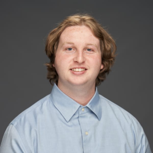 A young man with wavy, light brown hair and fair skin wearing a light blue button-up shirt poses in front of a plain gray background.