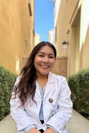 A person in a white coat sitting outdoors in a narrow pathway with plants and buildings in the background.