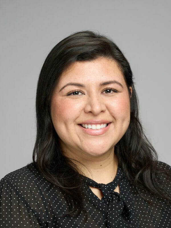 Woman with long dark hair and a black polka dot blouse smiles in front of a plain gray background.