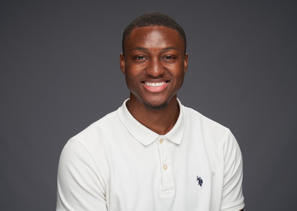 A man wearing a white polo shirt is smiling at the camera against a plain dark gray background.