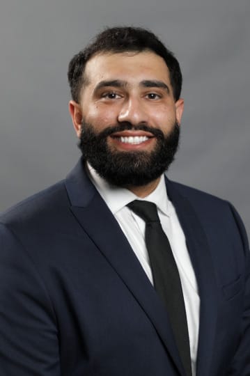 Man with a beard in a navy suit and black tie smiles against a gray background.