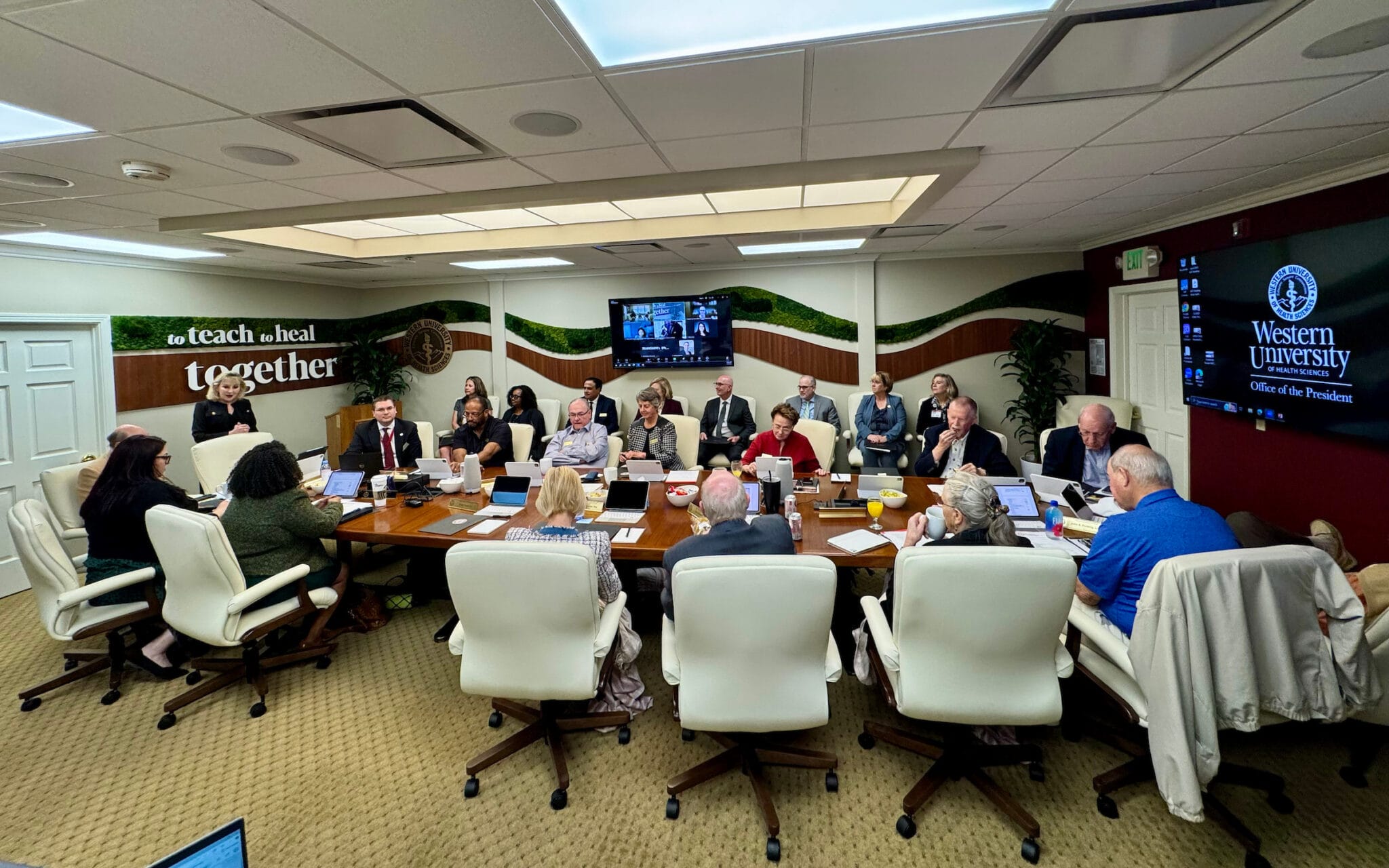 A group of people sit around a conference table in a meeting room, with screens displaying "Western University Office of the President" and a video call in the background.