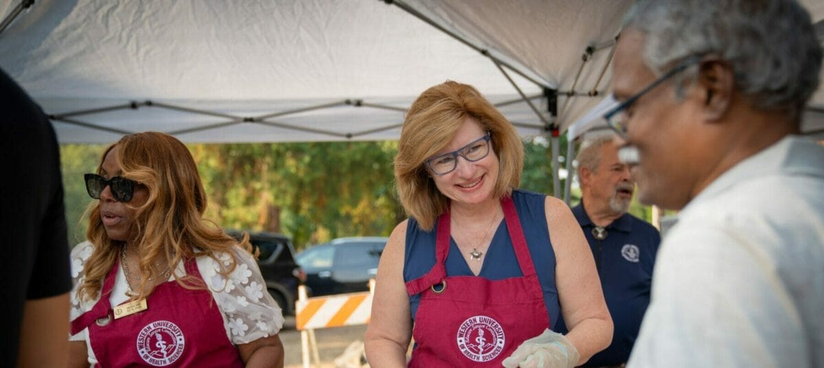 People wearing aprons and gloves serve food to others under a canopy at an outdoor gathering.