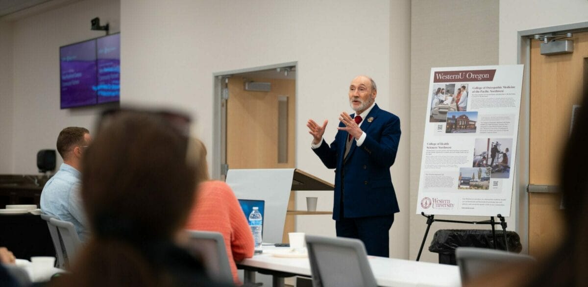 A man in a suit speaks at a podium with people seated at tables listening. A poster board beside him displays information and images.