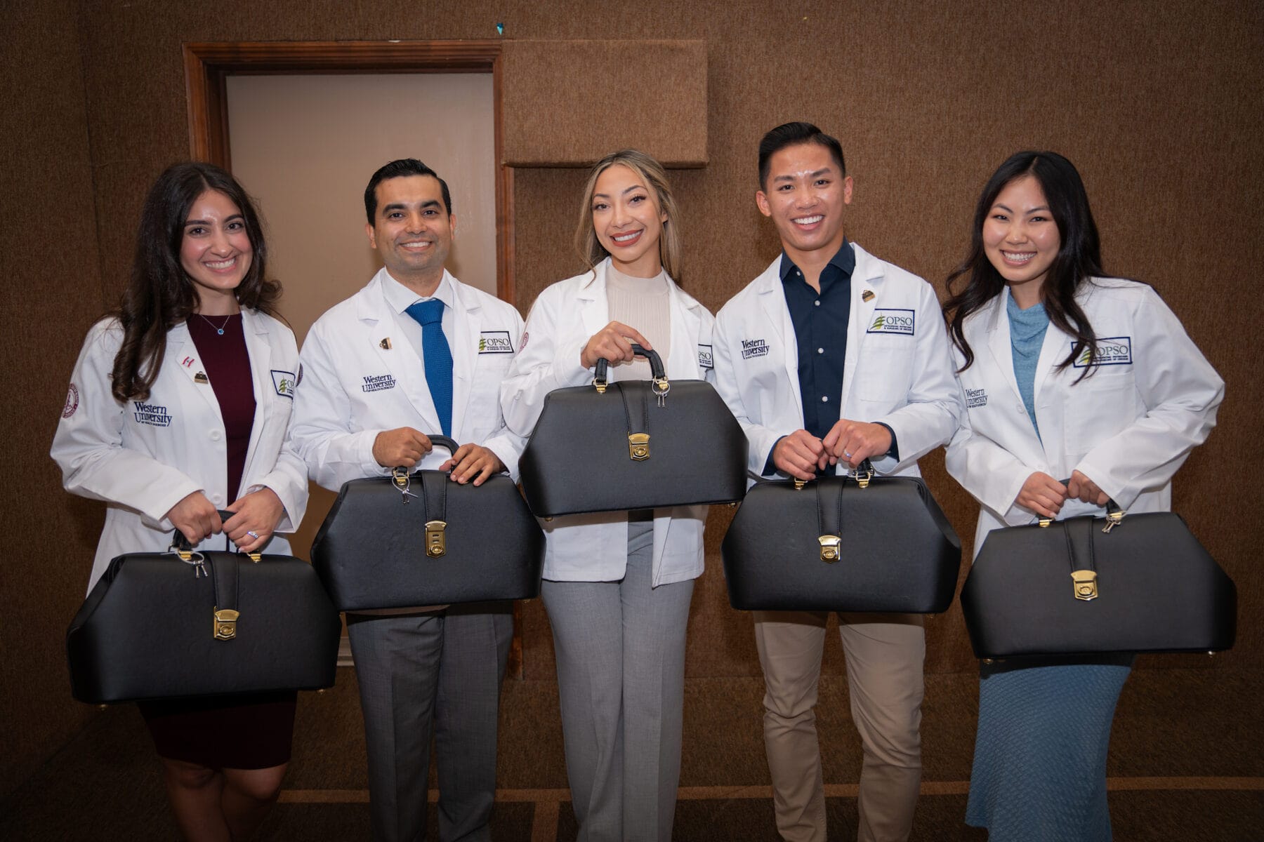 Five medical students in white coats hold black medical bags, smiling and standing in a row against a brown background.