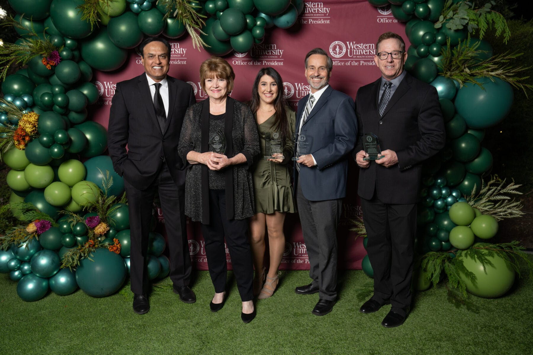 Five people in formal attire stand in front of a balloon arch and branded backdrop, holding glass awards and smiling for a group photo.