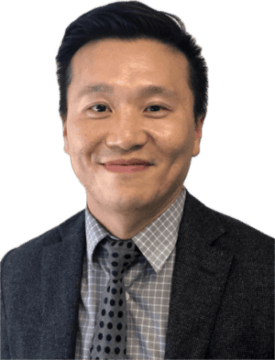 A man wearing a gray checkered shirt, a dark polka dot tie, and a gray suit jacket is smiling at the camera.
