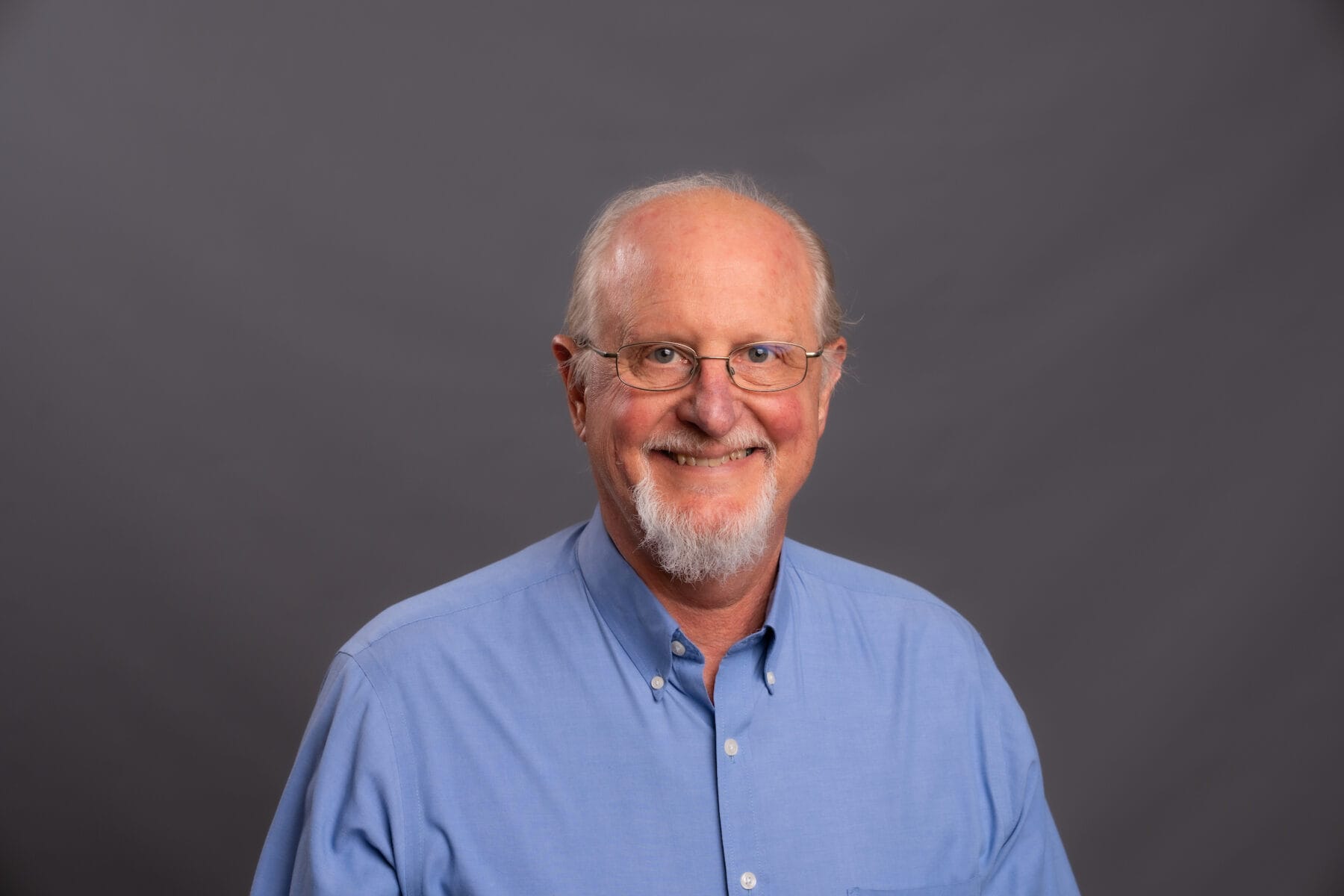 Older man with glasses and a white beard, wearing a light blue button-up shirt, smiling in front of a plain gray background.