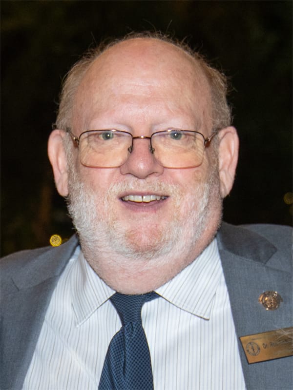 A man with glasses and a beard wearing a suit with a striped shirt and tie, smiling at the camera.