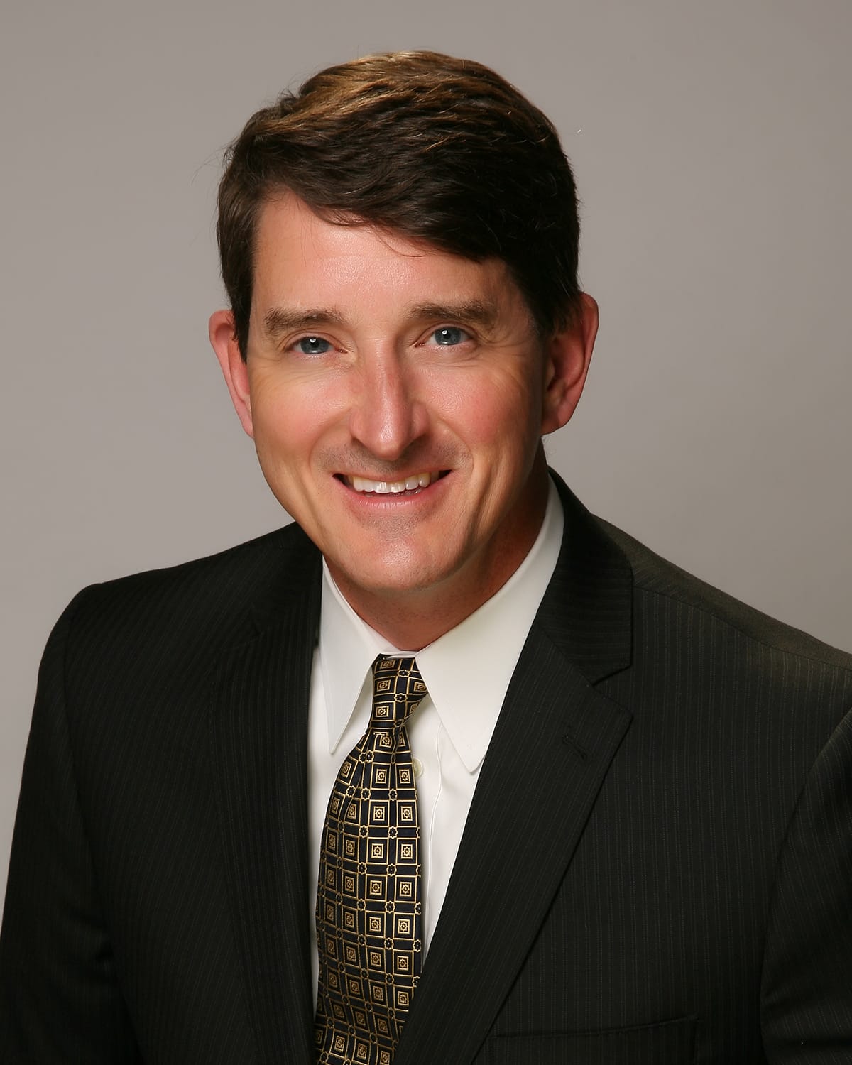 A man in a dark pinstripe suit, white shirt, and patterned tie poses for a professional studio portrait against a plain background.