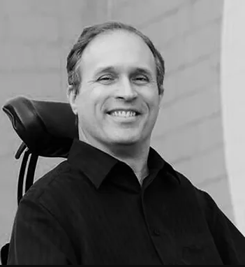 Man smiling in a black and white photo, seated in a wheelchair, wearing a dark shirt.