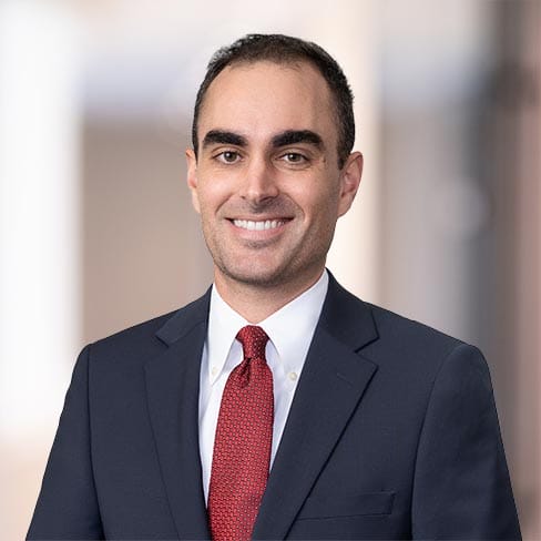 A man in a dark suit, white shirt, and red tie stands in front of a blurred indoor background, smiling at the camera.