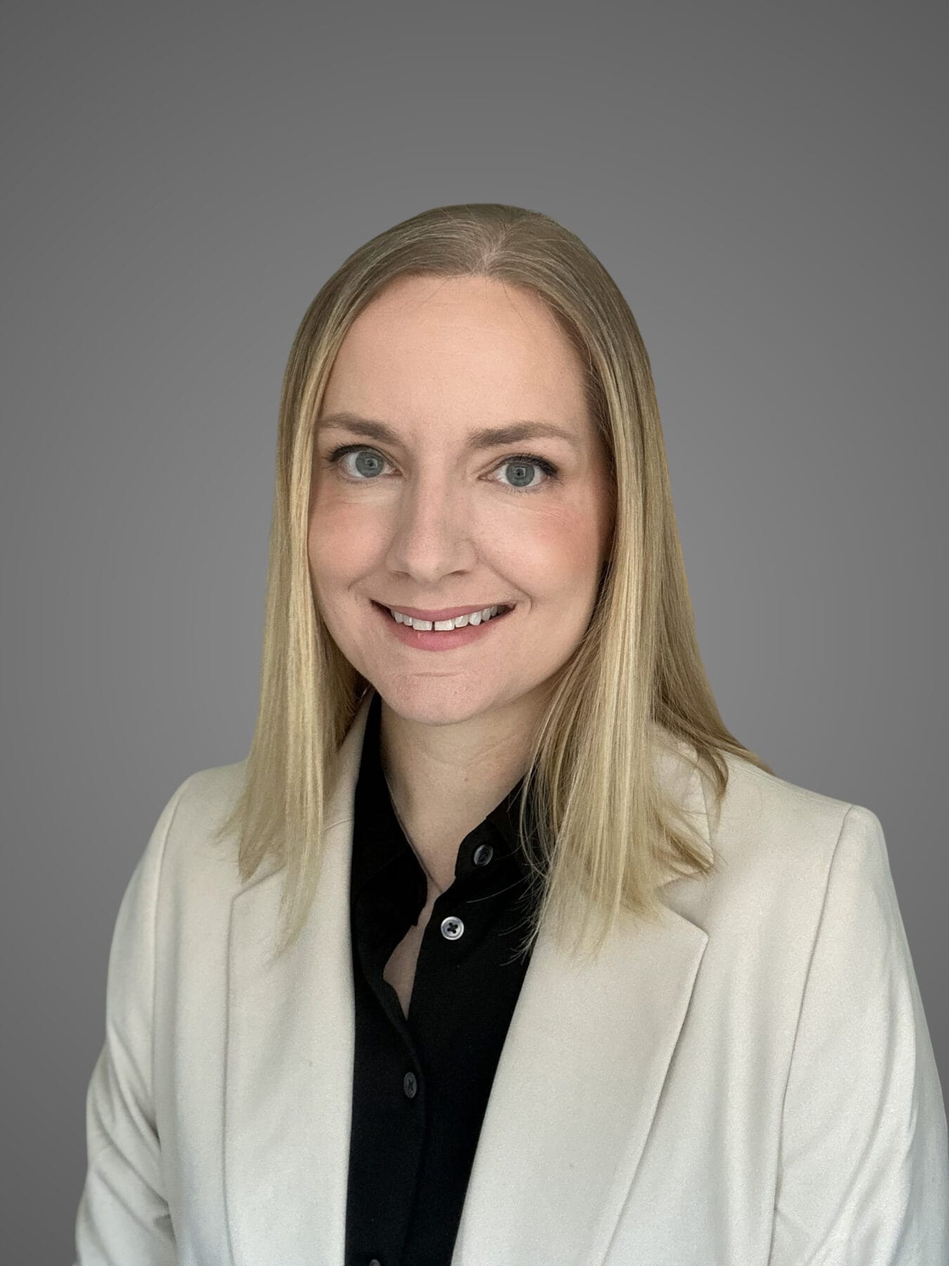 A woman with straight blonde hair, wearing a light-colored blazer over a black shirt, smiles at the camera against a plain gray background.