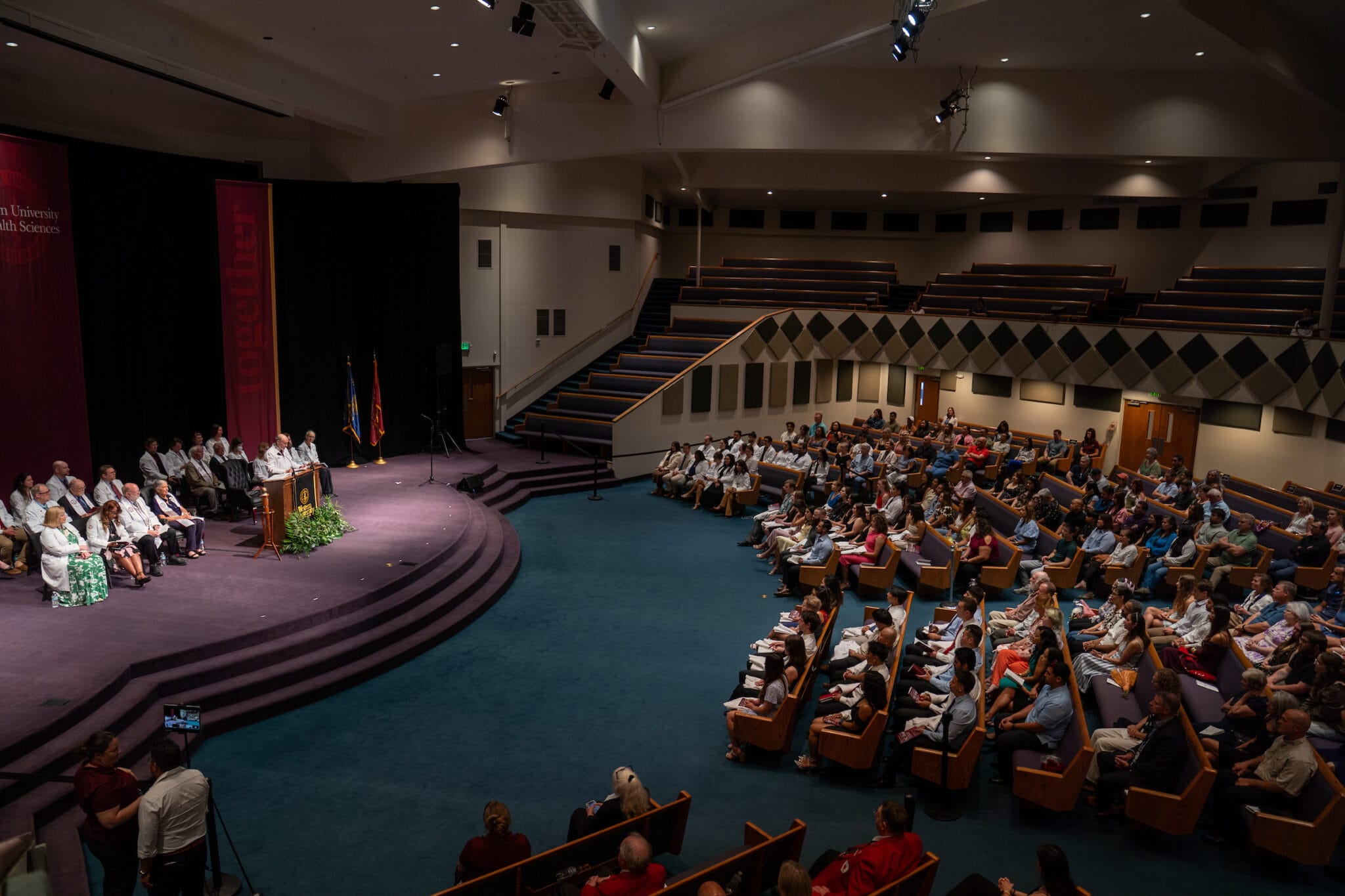 A speaker addresses an audience in a large auditorium with tiered seating; several people are seated on stage while attendees watch from the pews.