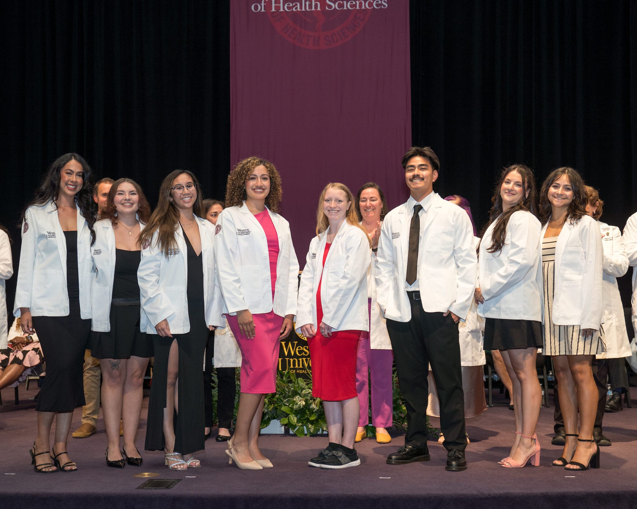A group of eight people in white coats pose and smile on stage at a ceremony, with a purple curtain and university logo in the background.