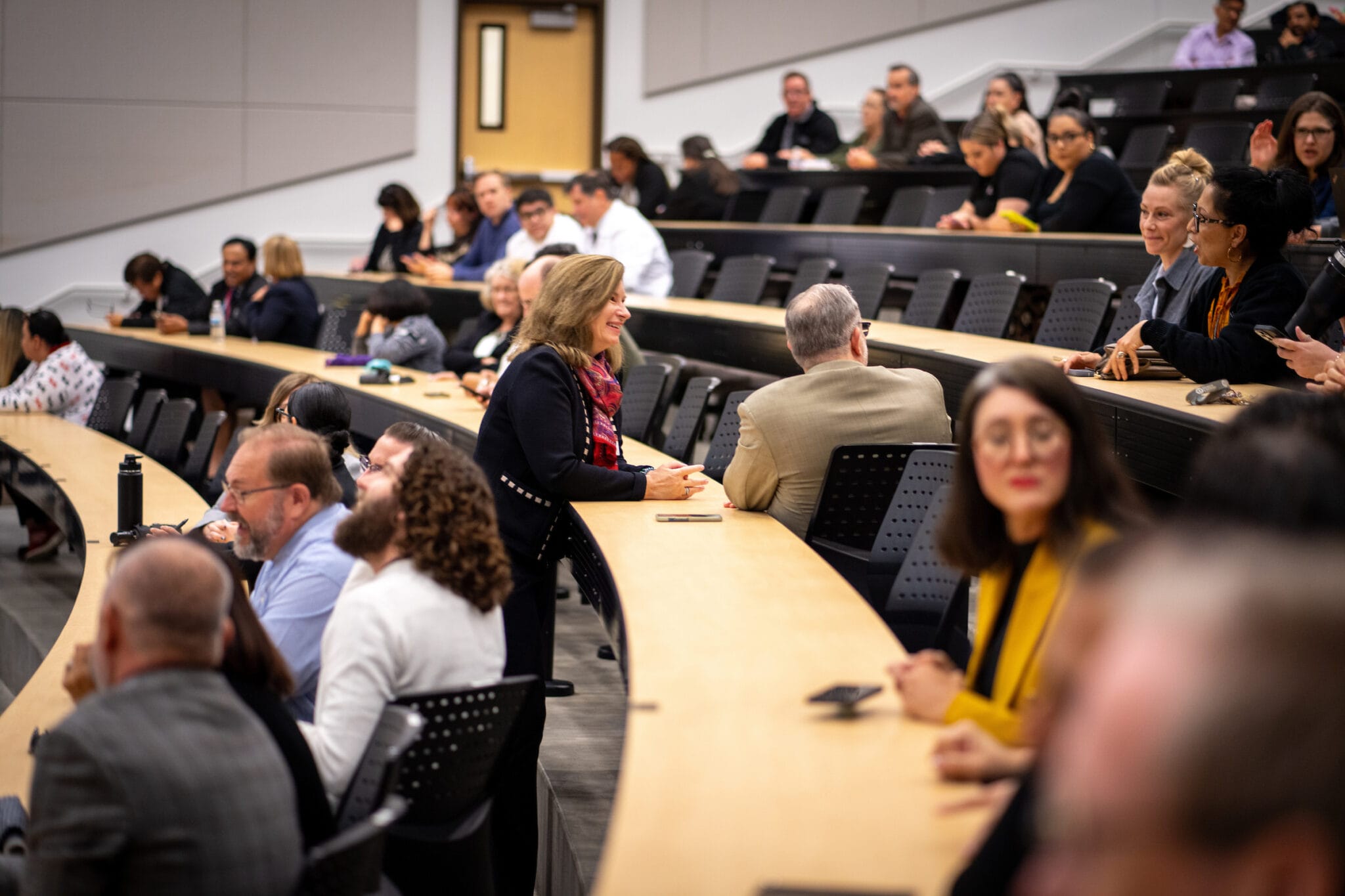 People seated in a modern lecture hall, some conversing and others waiting, with a focus on a woman standing and talking to a man.