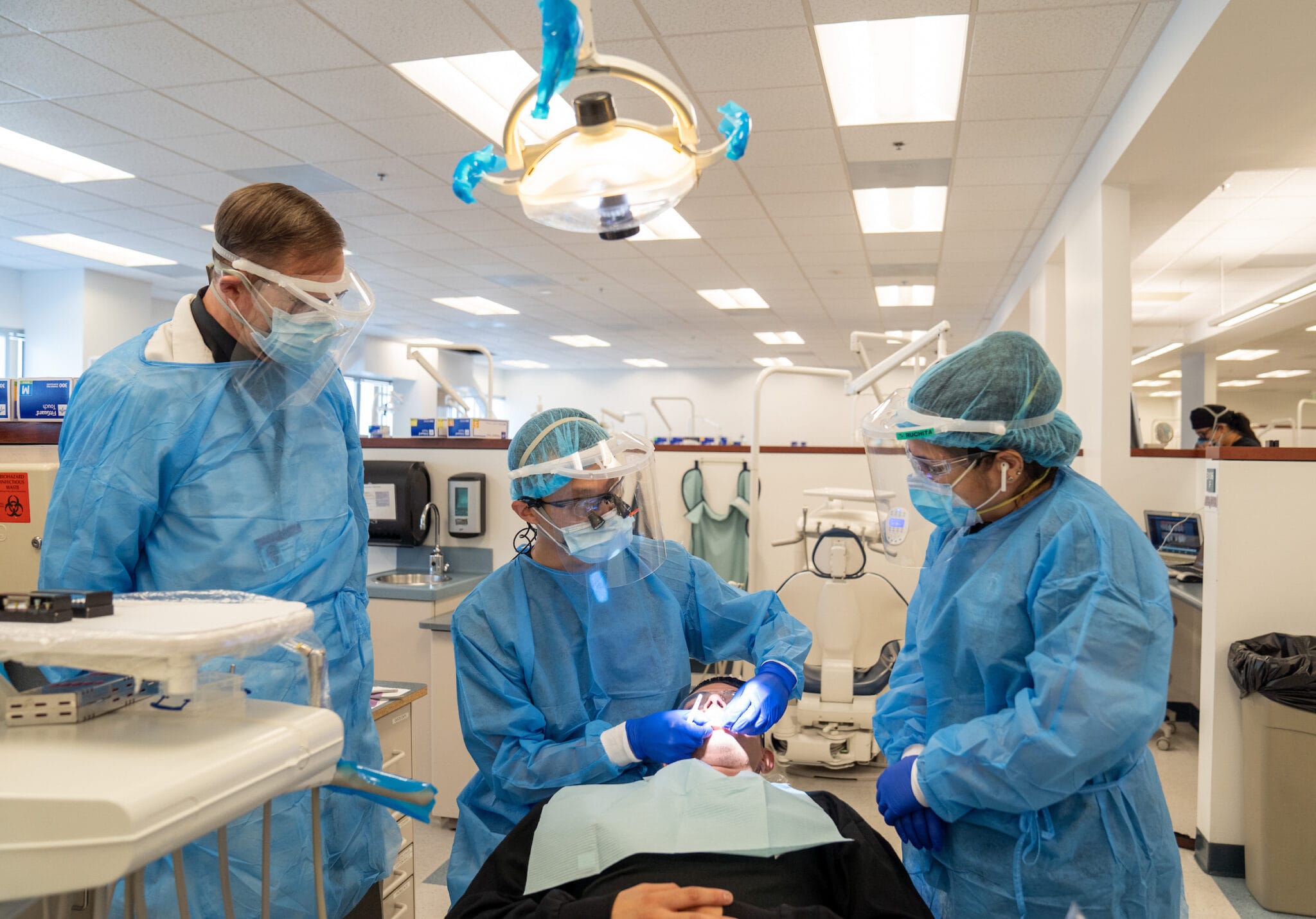 Three dental professionals in blue protective gowns and face shields perform a procedure on a patient in a brightly lit dental clinic.