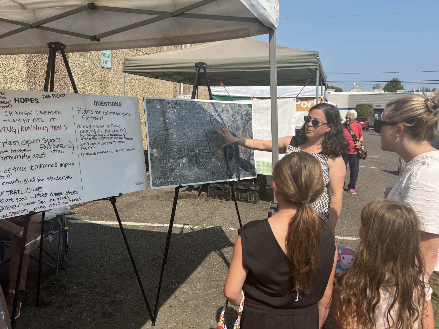 A woman points to a map on an easel, explaining to three individuals at an outdoor event with informational boards and tents in the background.