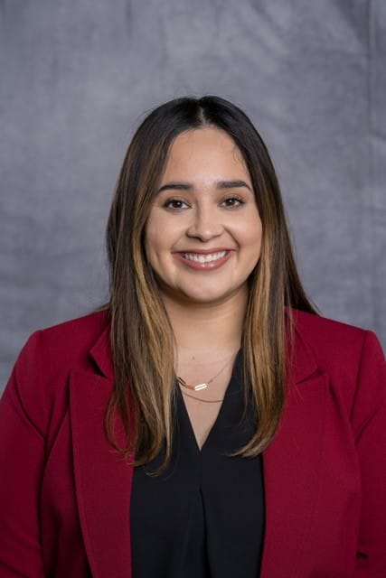 A woman with long brown hair wearing a red blazer and black top smiles in front of a gray backdrop.