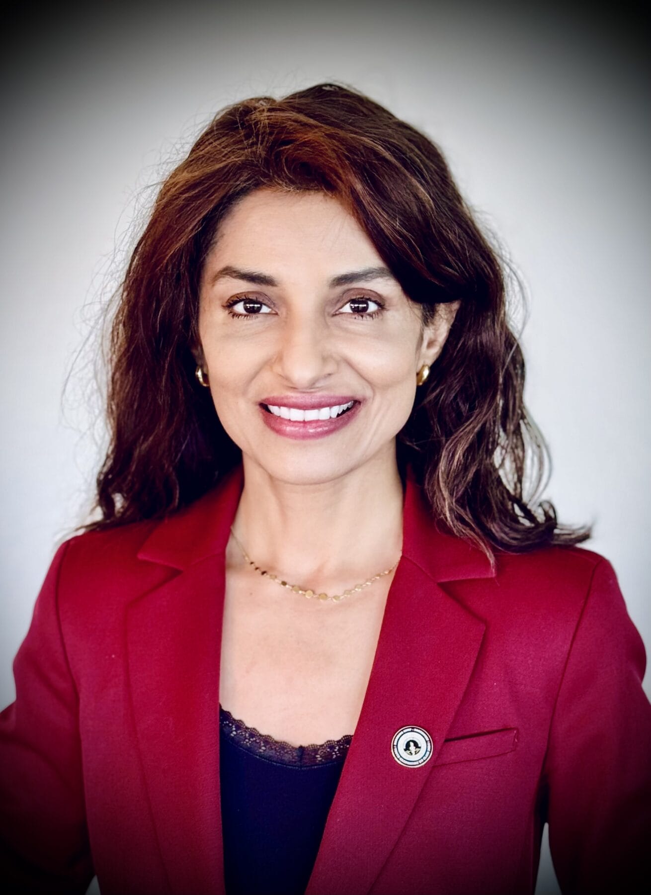 Woman with long brown hair wearing a red blazer, black top, and a pin, smiling at the camera against a plain light background.