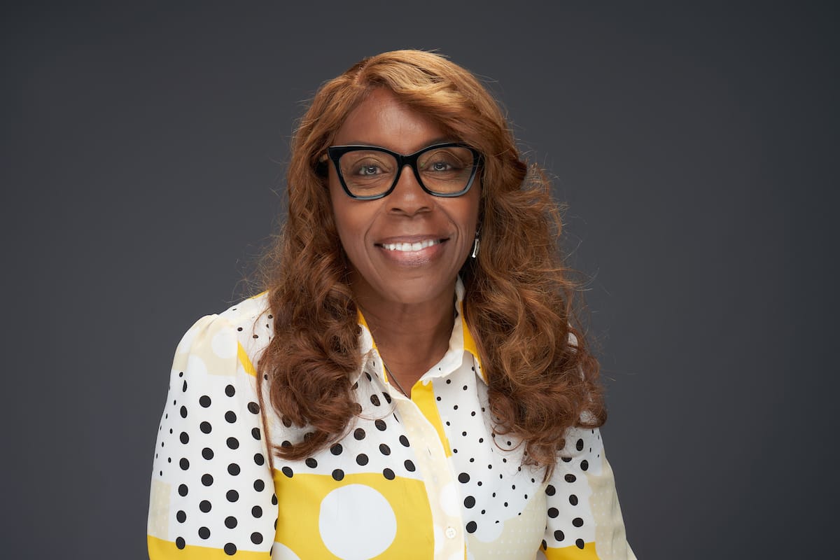 A person with curly, reddish-brown hair, wearing glasses and a white blouse with a yellow and black polka dot pattern, smiles in front of a neutral background.