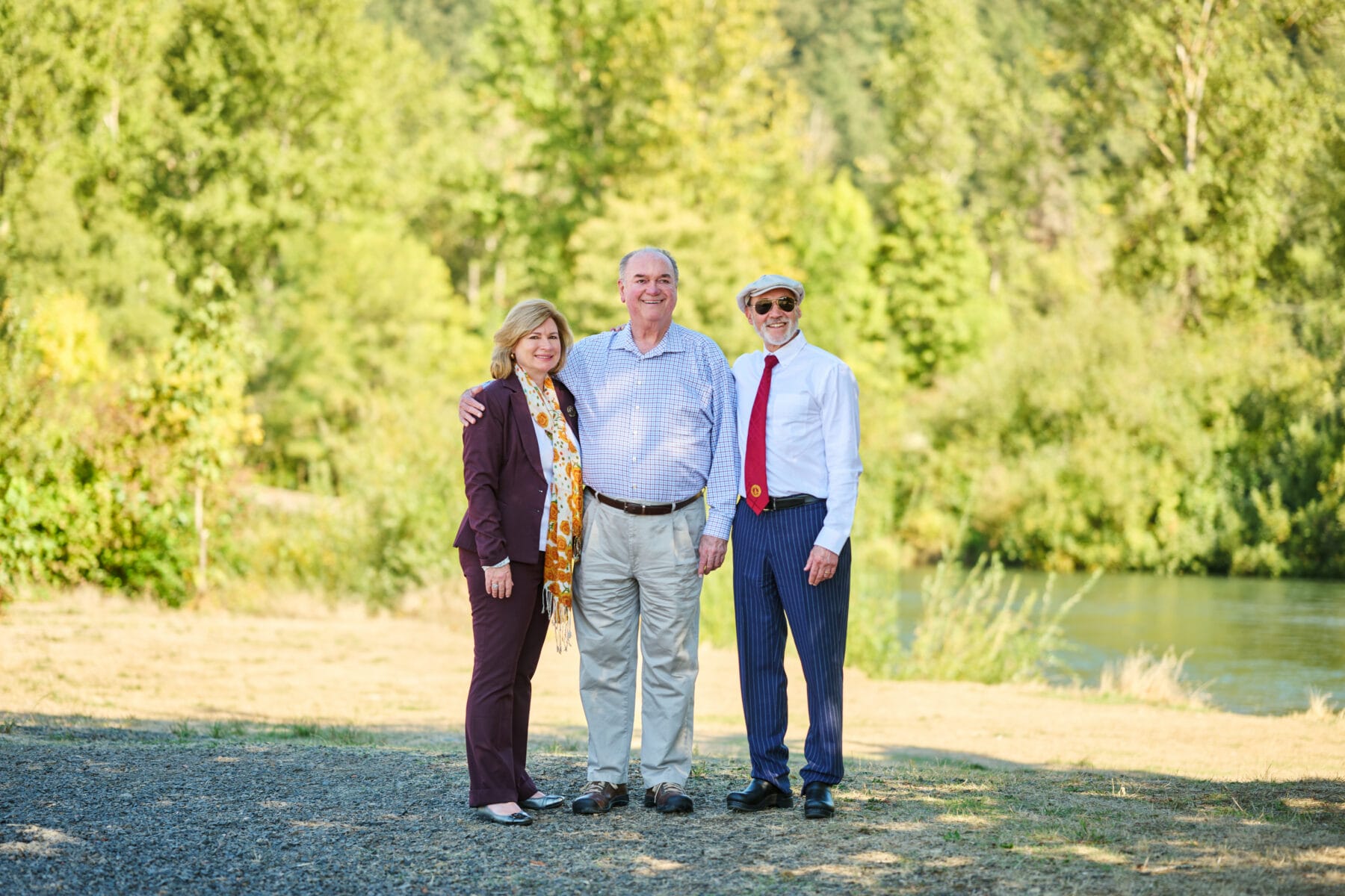 Three people standing outdoors near a body of water and trees, with two men in dress shirts and one woman in a suit, posing for a photo.