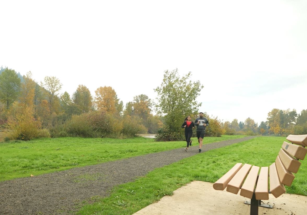 Two people jog on a gravel path near a wooden bench in a park with green grass and autumn trees in the background.