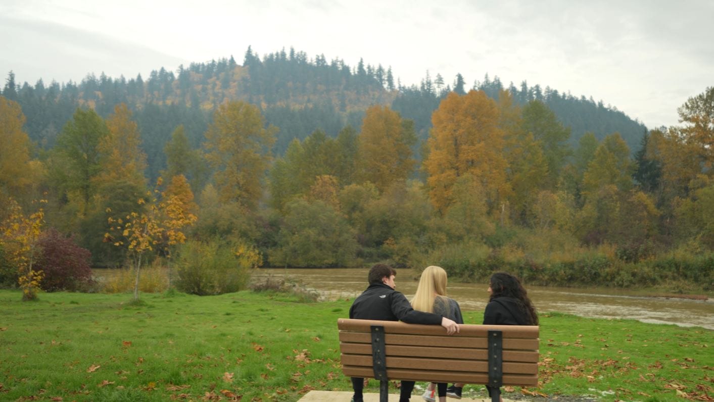 Three people sit on a bench facing a river with a backdrop of autumn trees and hills.