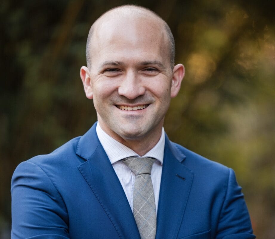 A man in a blue suit and tie stands outdoors with his arms crossed, smiling at the camera.